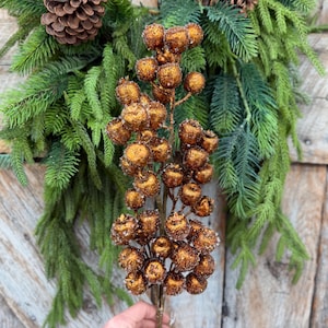 May include: A decorative floral stem featuring numerous round, metallic copper-colored berries with a textured, glittery finish. The stem is positioned against a background of green pine needles and a brown pinecone, creating a festive appearance.