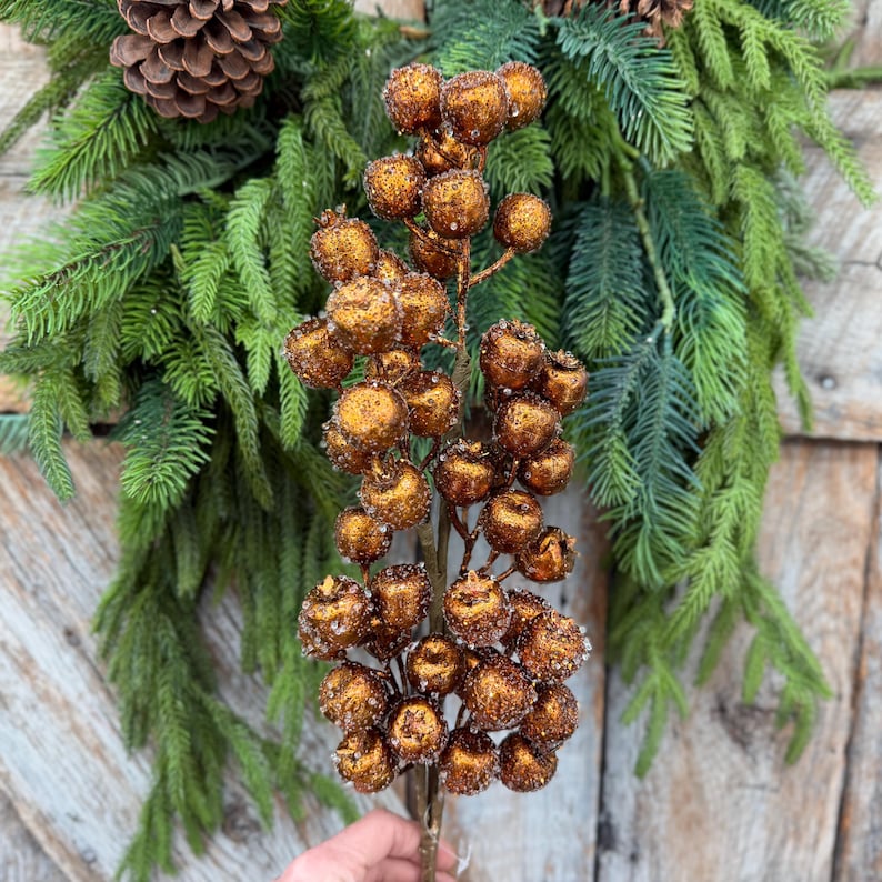 May include: A decorative floral stem featuring numerous round, metallic copper-colored berries with a textured, glittery finish. The stem is positioned against a background of green pine needles and a brown pinecone, creating a festive appearance.