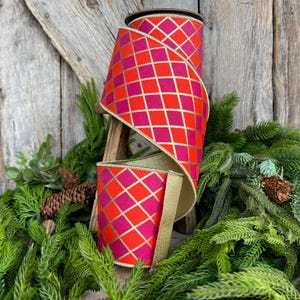 May include: A decorative ribbon roll with a vibrant pattern of red and fuchsia diamonds outlined in gold. The ribbon is displayed on a wooden stand, surrounded by greenery and pinecones, suggesting a festive or seasonal theme.