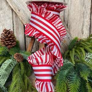 May include: A spool of red and white striped ribbon with ruffled edges. The ribbon is displayed with pinecones and evergreen branches, suggesting a holiday or seasonal theme. The ribbon's design features bold, vertical stripes.