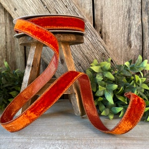 May include: A spool of burnt orange velvet ribbon with a satin edge. The ribbon is wrapped around a wooden stool and is laying on a wooden surface.