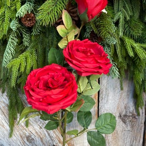 May include: A vibrant red rose stem with two fully bloomed roses and a bud, surrounded by green leaves. The roses are set against a backdrop of evergreen branches and pinecones, suggesting a festive or decorative theme.