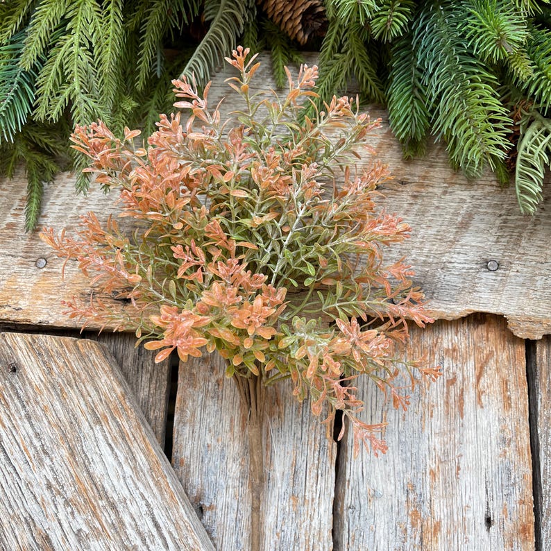 May include: A bundle of artificial greenery with orange and green leaves. The faux plant is set against a rustic wooden background, with evergreen branches and pinecones in the background.