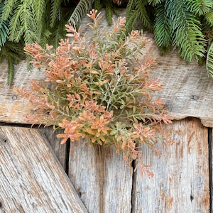 May include: A bundle of artificial greenery with orange and green leaves. The faux plant is set against a rustic wooden background, with evergreen branches in the top corners.