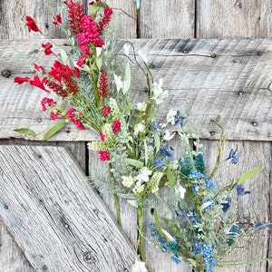 May include: Three artificial flower stems with red, white, and blue flowers. The stems are arranged in a bouquet against a weathered wooden background.