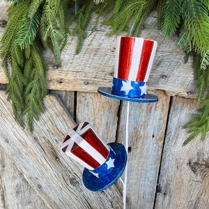 May include: Two red, white, and blue patriotic top hats with stars on a blue background. The hats are on white sticks and are leaning against a weathered wooden fence.