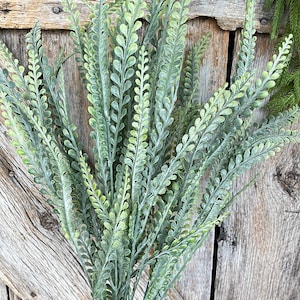 May include: A bundle of artificial greenery with small, rounded leaves in varying shades of green. The stems are a darker green, and the leaves have a slightly frosted appearance. The background is a rustic wooden surface.