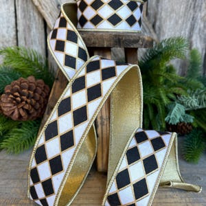 May include: A roll of decorative ribbon with a black and white diamond pattern. The ribbon has a gold metallic backing and gold trim. The ribbon is displayed on a wooden surface with pine needles and a pine cone in the background.