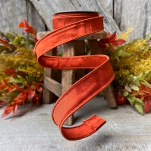 May include: A spool of burnt orange velvet ribbon with a stitched edge. The ribbon is wrapped around a wooden stool.
