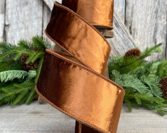 May include: A roll of wide, bronze-coloured ribbon with a satin finish. The ribbon has a brown stitched edge and is partially unrolled, revealing its texture. The background includes pine branches and a wooden surface, suggesting a holiday or craft theme.