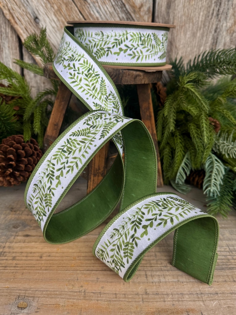 May include: A spool of wired ribbon featuring a white background with a repeating green watercolor fern leaf pattern. The ribbon has a solid green underside and finished edges, shown unspooled in a decorative loop on a wooden surface.