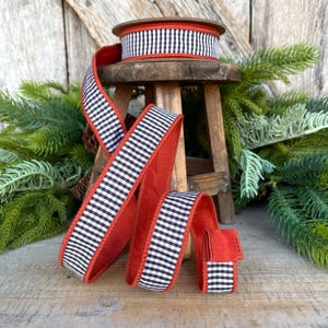 May include: A spool of black and white checkered ribbon with a red backing sits atop a small wooden stool. The ribbon is unraveled, displaying its pattern and color. Green pine branches and a wooden background complete the scene.