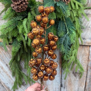 May include: A decorative floral stem featuring numerous round, metallic copper-colored berries with a textured, glittery finish. The stem is positioned in front of a lush arrangement of green pine needles and a brown pinecone.