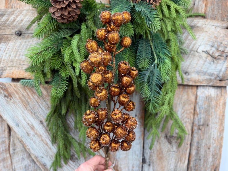 May include: A decorative floral stem featuring numerous round, metallic copper-colored berries with a textured, glittery finish. The stem is positioned in front of a lush arrangement of green pine needles and a brown pinecone.