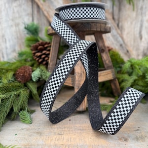May include: A roll of wired ribbon featuring a black and white checkered pattern. The edges of the ribbon are finished with a black glittery texture that sparkles, providing a distinct contrast to the central geometric square design.