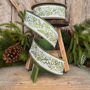 May include: A spool of wired ribbon featuring a white background with a repeating green watercolor fern leaf pattern. The ribbon has a solid green underside and finished edges, shown coiled and draped in a spiral shape to display the print.
