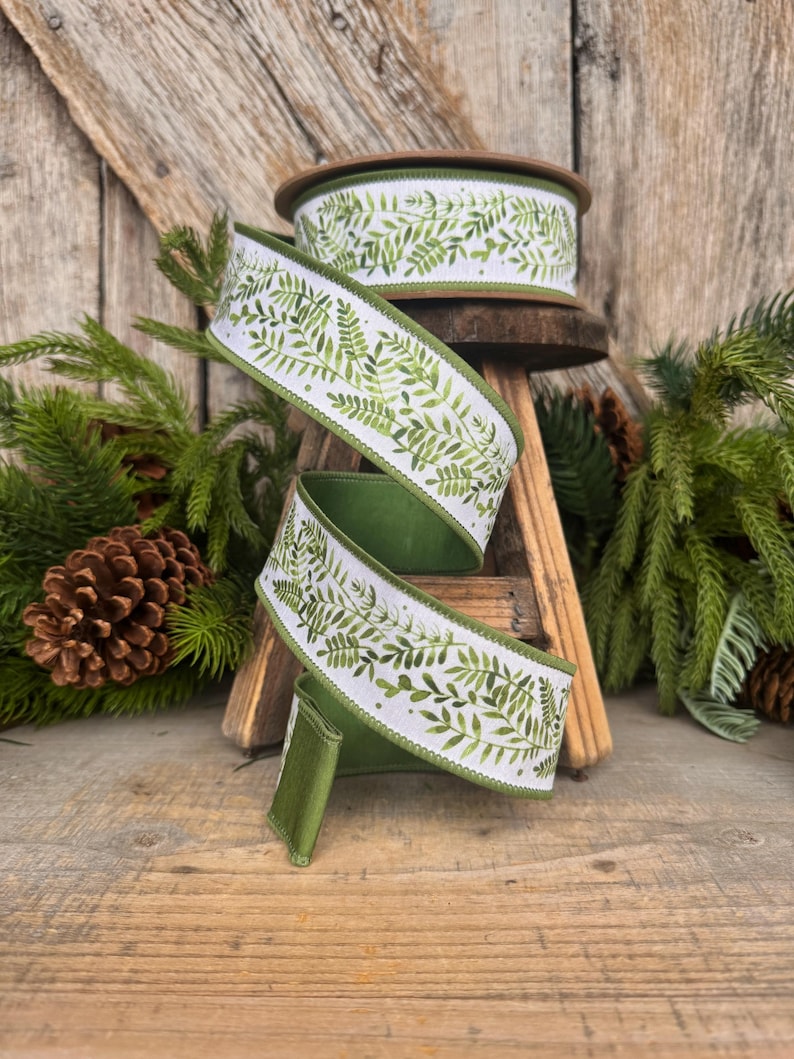 May include: A spool of wired ribbon featuring a white background with a repeating green watercolor fern leaf pattern. The ribbon has a solid green underside and finished edges, shown coiled and draped in a spiral shape to display the print.