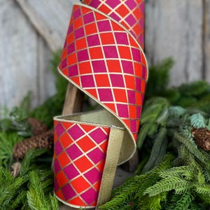 May include: A roll of decorative ribbon with a vibrant harlequin pattern in shades of red and fuchsia, accented with gold lines and trim. The ribbon is displayed against a backdrop of greenery, suggesting its use for holiday or festive decor.