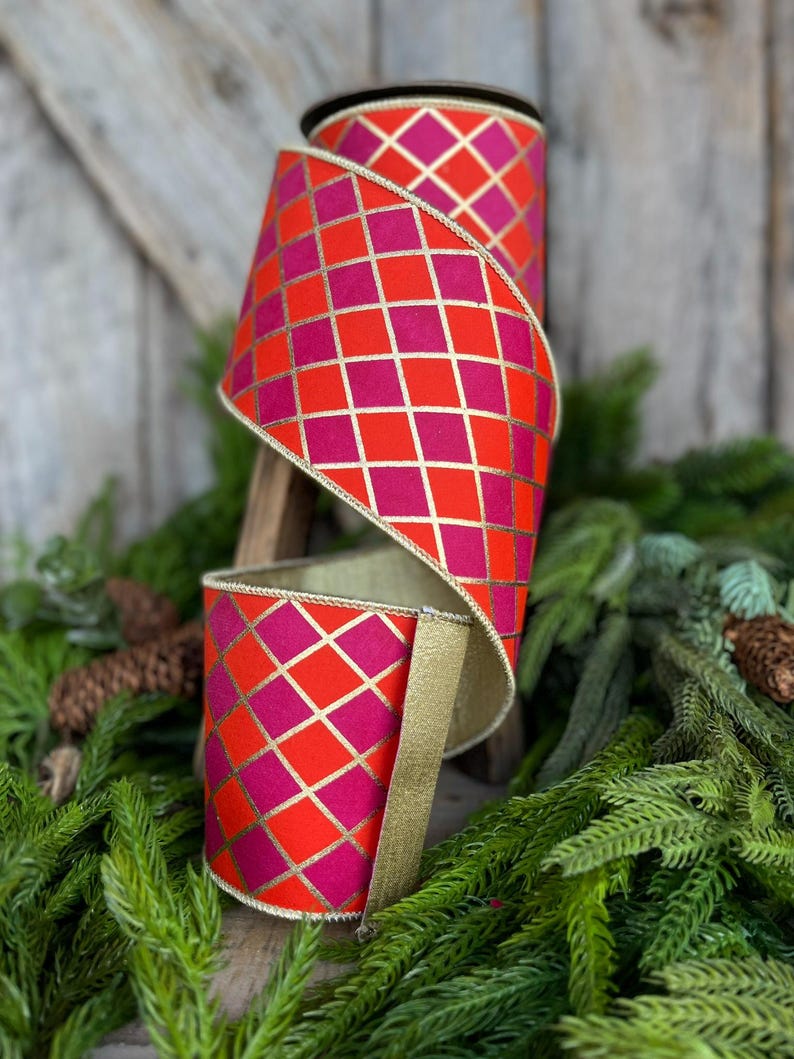 May include: A roll of decorative ribbon with a vibrant harlequin pattern in shades of red and fuchsia, accented with gold lines and trim. The ribbon is displayed against a backdrop of greenery, suggesting its use for holiday or festive decor.