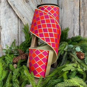 May include: A roll of decorative ribbon with a vibrant red and pink diamond pattern, accented with gold lines. The ribbon is displayed on a wooden stand, surrounded by greenery and pinecones, suggesting a festive or holiday theme.