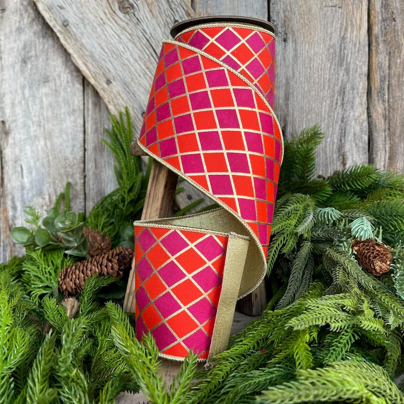 May include: A roll of decorative ribbon with a vibrant red and pink diamond pattern, accented with gold lines. The ribbon is displayed on a wooden stand, surrounded by greenery and pinecones, suggesting a festive or holiday theme.