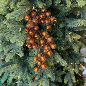 May include: A decorative branch featuring numerous round, copper-colored berries with a textured, glittery finish. The berries are clustered together on thin stems, creating a dense, hanging arrangement against a background of green pine needles.