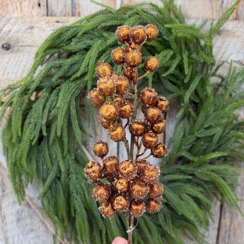 May include: A decorative floral stem featuring numerous round, copper-colored berries with a textured, glittery finish. The berries are clustered along a thin, brown branch, creating a dense, spherical arrangement of metallic-toned fruit.