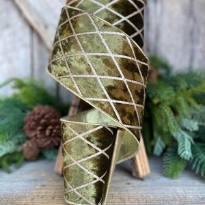May include: A roll of olive green velvet ribbon with a gold diamond pattern. The ribbon is displayed on a wooden spool, with a rustic wooden background and evergreen accents.