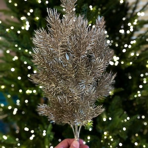 May include: A silver-colored artificial pine sprig with a glittery finish. The sprig is held in a hand against a blurred background of a Christmas tree with white lights.