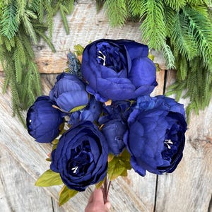 May include: A bouquet of deep blue artificial peonies with green leaves. The flowers are held in a hand against a rustic wooden background, with green foliage in the upper corners.
