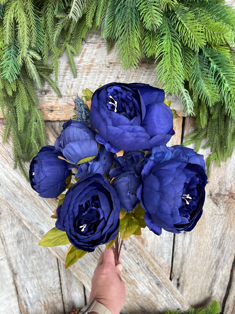 May include: A bouquet of deep blue artificial peonies with green leaves. The flowers are held in a hand against a rustic wooden background, with green foliage in the upper corners.