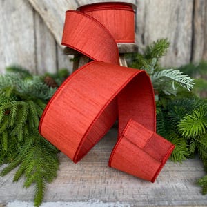 May include: A roll of red ribbon with a textured surface and a contrasting red edge. The ribbon is displayed with a portion unrolled, showcasing its width and texture. The background includes greenery and a wooden surface, suggesting a craft or holiday theme.