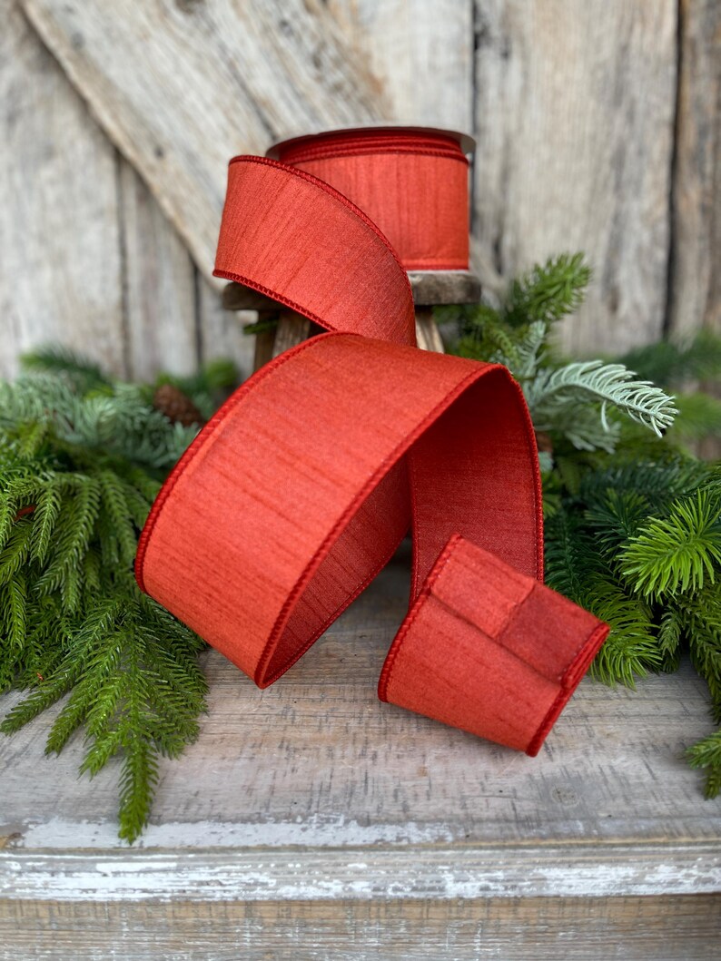 May include: A roll of red ribbon with a textured surface and a contrasting red edge. The ribbon is displayed with a portion unrolled, showcasing its width and texture. The background includes greenery and a wooden surface, suggesting a craft or holiday theme.
