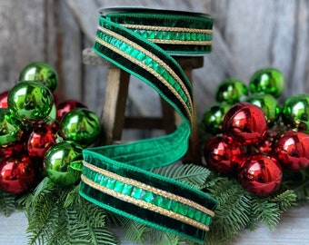 May include: A spool of emerald green velvet ribbon with gold and jewel accents. The ribbon is displayed with red and green Christmas ornaments and evergreen sprigs, suggesting holiday decor.