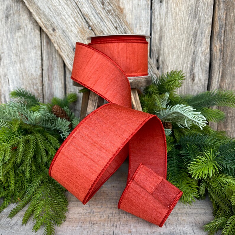 May include: A roll of wide, textured, red ribbon with a matching red trim. The ribbon is displayed with a portion unrolled, revealing its texture and color. The background includes greenery and a rustic wooden surface, suggesting a holiday or craft theme.