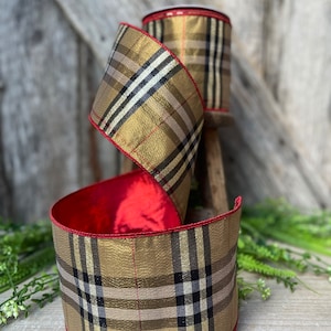 May include: A roll of decorative ribbon with a gold and black plaid pattern. The ribbon has a red satin lining and red trim. The ribbon is displayed on a wooden surface with greenery in the background.
