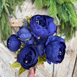 May include: A bouquet of deep blue peonies with white accents, held against a rustic wooden background. The flowers have multiple layers of petals and are surrounded by green leaves. The arrangement is accented by a backdrop of green pine branches.