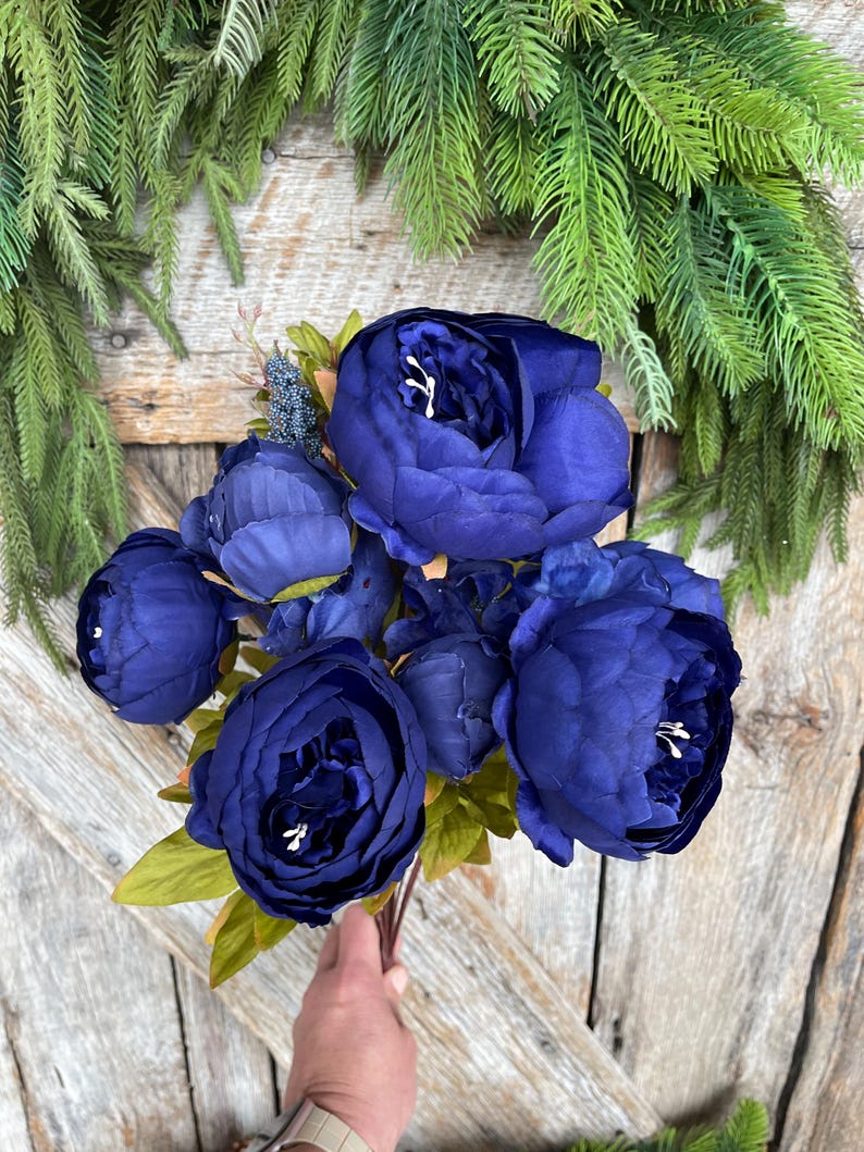 May include: A bouquet of deep blue peonies with white accents, held against a rustic wooden background. The flowers have multiple layers of petals and are surrounded by green leaves. The arrangement is accented by a backdrop of green pine branches.