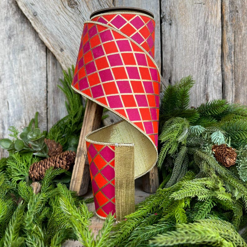May include: A decorative ribbon roll with a red and pink diamond pattern, accented with gold trim. The ribbon is displayed on a wooden stand, surrounded by greenery and pinecones, suggesting a holiday or craft theme.