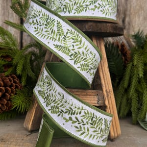 May include: A roll of white wired ribbon featuring a repeating green fern leaf pattern. The ribbon has solid green finished edges and is coiled in a spiral shape, displaying the botanical print clearly against the white fabric background.