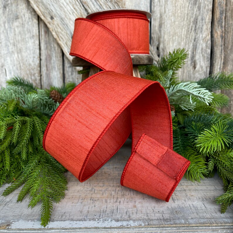 May include: A roll of wide, red ribbon with a textured appearance and a darker red trim. The ribbon is displayed with a portion unrolled, showcasing its texture and color. The background includes greenery and a rustic wooden surface, suggesting a holiday or craft theme.