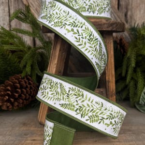 May include: A spool of white ribbon featuring a repeating green watercolor fern leaf pattern. The ribbon has a solid green fabric backing and finished stitched edges, shown spiraling downward from a wooden spool.
