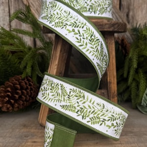 May include: A spool of white ribbon featuring a repeating green watercolor fern leaf pattern. The ribbon has a solid green fabric backing and finished stitched edges, shown spiraling downward from a wooden spool.