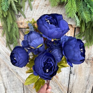 May include: A bouquet of deep blue artificial peonies with green leaves. The flowers are in full bloom, with multiple petals visible. The bouquet is held against a wooden background, with green foliage in the background.