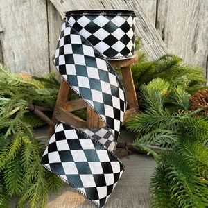 May include: A roll of decorative ribbon with a black and white harlequin pattern. The ribbon is displayed on a small wooden stool, with evergreen branches and pinecones in the background. The ribbon is approximately 2.5 inches wide.