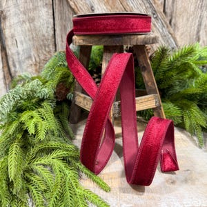 May include: A spool of deep red velvet ribbon with a soft, textured pile and finished edges. The ribbon is draped in loose folds, showcasing its rich, saturated color and light-reflecting surface, creating a continuous, elegant, and flexible fabric strip.