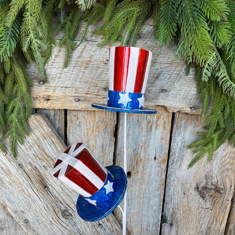 May include: Two red, white, and blue patriotic top hats with white stars on a blue band. The hats are on white sticks against a wooden background.