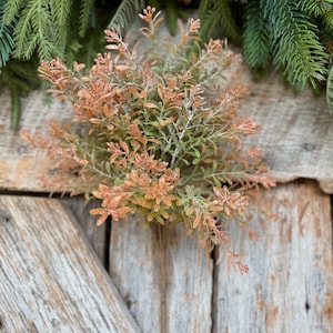 May include: A close-up of a decorative floral arrangement featuring a cluster of small, orange and green leaves. The arrangement is set against a rustic wooden background, with evergreen branches and pine cones in the background.