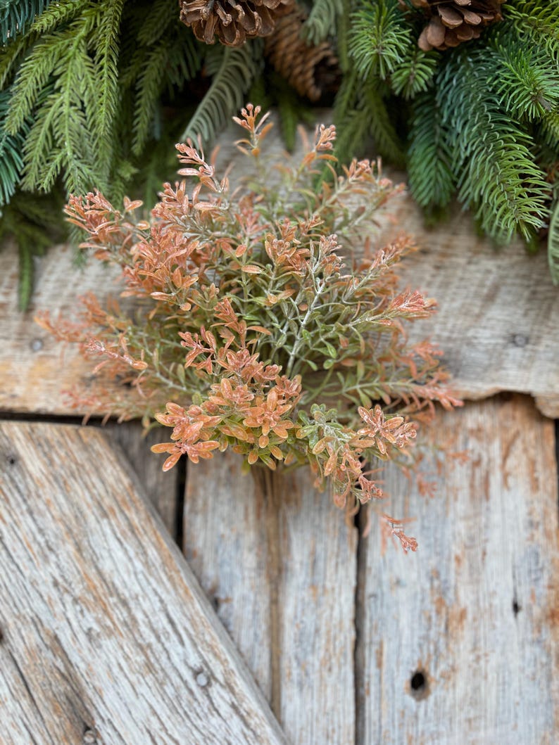 May include: A close-up of a decorative floral arrangement featuring a cluster of small, orange and green leaves. The arrangement is set against a rustic wooden background, with evergreen branches and pine cones in the background.