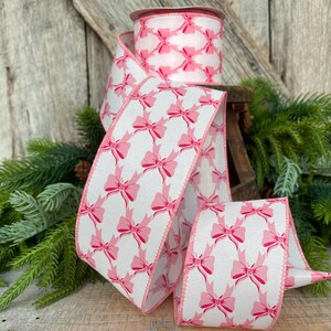 Peut inclure: Un rouleau de ruban blanc avec un motif r&eacute;p&eacute;t&eacute; de n&oelig;uds roses. Le ruban a un bord cousu rose et est pr&eacute;sent&eacute; avec de la verdure et un fond en bois. Les n&oelig;uds sont espac&eacute;s uniform&eacute;ment sur le ruban.